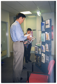 COL photo of three people (one obscured) standing in front of a book display. The books are contained inside clear perspex holders, which have been affixed to a freestanding display board. Each person is looking down at an open book, which they are holding in their hands.