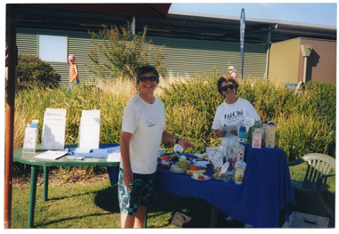 COL photo of two women standing on either side of a table covered in a bright blue tablecloth. One of the women is wearing a t-shirt printed with 'Tai Chi for ARTHRITIS by Dr Paul Lam' with the Arthritis Foundation of Victoria logo and some additional text 'supported by Arthritis Foundation' (partially obscured) below. On top of the table, there are various fresh and packaged food items, which have been labelled. There is a second table beside this one, containing various factsheets and leaflets on small display stands. Behind them is a building with a corrugated iron feature wall.