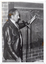 B&W photo of a man standing next to a blackboard, which has been affixed to a brick wall. He is speaking into a microphone on a stand while pointing to the blackboard. On the blackboard, the following text is written: Assessmt - Med -Phys -Psycho Soc