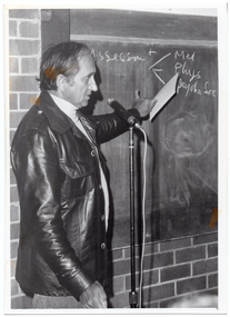 B&W photo of a man standing next to a blackboard, which has been affixed to a brick wall. He is speaking into a microphone on a stand while pointing to the blackboard. On the blackboard, the following text is written: Assessmt - Med -Phys -Psycho Soc