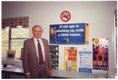 COL photo of a man standing in front of a row of tables set against the wall. The wall is covered with various posters, and there is a window on either side. Poster titles include: "If old age is catching up, walk a little faster" (VicHealth, Active At Any Age); "Table tennis [keeps] you moving. Move it or lose it"; "Ben Back-Bender says WATCH YOUR BACK"; "The WALK of life. Well-being Aerobic exercise Life Extension Kilograms" (Institute of Health & Fitness); "Betty Bones [illegible] YOU CAN [illegible] BONES STRONG! MOVE IT OR LOSE IT".
