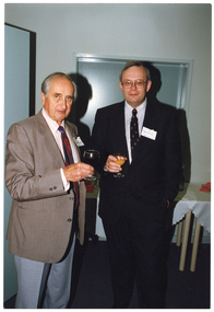 COL photo of two men standing indoors, near a doorway. On the wall behind them is a butler's servery, which is closed.