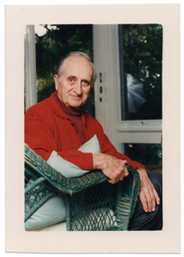 COL photo of a man sitting in a green wicker chair. He has one arm pressing down on top of a cushion, which is placed against the arm of the chair. Behind him are some glass doors and windows, through which there is a view of several trees in a garden.