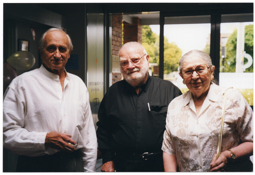 COL photo of three people standing in the foyer of a building. In the background, tall trees and a multistorey building (partially obscured) are visible through the large glass front doors and windows.