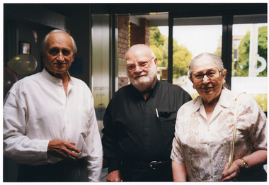 COL photo of three people standing in the foyer of a building. In the background, tall trees and a multistorey building (partially obscured) are visible through the large glass front doors and windows.