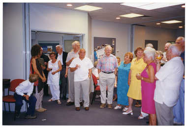 COL photo of a large group of people, some standing and some seated, gathered in a large meeting room. Some people are holding a glass in one hand (filled with either wine, orange juice or water). In the background, there is a set of bi-fold doors against one wall which have been folded back into the open position. There is also a display board with Photographs and posters along the same wall.