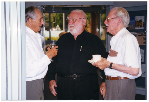 COL photo of three men standing in a foyer, chatting. One of them is holding a glass of red wine while another is holding a cup and saucer. In the background, there is a view of some trees through a set of glass doors.
