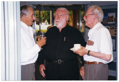 COL photo of three men standing in a foyer, chatting. One of them is holding a glass of red wine while another is holding a cup and saucer. In the background, there is a view of some trees through a set of glass doors.