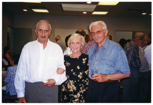 COL photo of three people standing in a large room, posing for a photo. Behind them are groups of people, both standing and seated at tables.