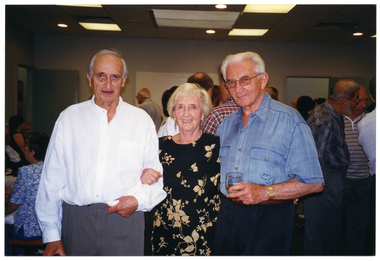 COL photo of three people standing in a large room, posing for a photo. Behind them are groups of people, both standing and seated at tables.
