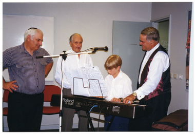 COL photo of a boy playing a Roland E-500 electric keyboard. Standing around him are three men. One of them is wearing a 'kippah' or 'yarmulke' - a small hat traditionally worn by Jewish boys and men. There is a microphone on a stand poised over the keyboard, above the boy's head.
