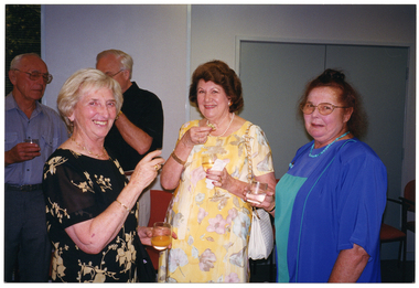 COL photo of three women, standing in a large room, posing for a photo. They are holding drinks and/or finger food in their hands. In the background, there are two men chatting.