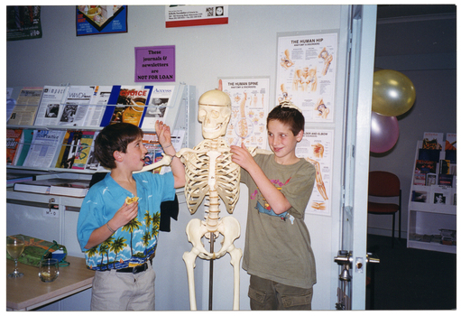 COL photo of two children posing with a skeleton on a vertical stand. Behind them, above some filing cabinets, are some shelves containing printed resources. On the wall, there is a sign above the shelves that says "These journals & newsletters are NOT FOR LOAN". There are also several posters on the wall, including titles such as "THE HUMAN SPINE", "THE HUMAN HIP", and "...LDER AND ELBOW" (partially obscured). On a table in front of the filing cabinets, there is a glass of wine and another glass with a drink. Through a doorway, two balloons are visible.