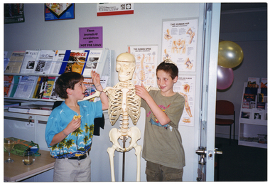 COL photo of two children posing with a skeleton on a vertical stand. Behind them, above some filing cabinets, are some shelves containing printed resources. On the wall, there is a sign above the shelves that says "These journals & newsletters are NOT FOR LOAN". There are also several posters on the wall, including titles such as "THE HUMAN SPINE", "THE HUMAN HIP", and "...LDER AND ELBOW" (partially obscured). On a table in front of the filing cabinets, there is a glass of wine and another glass with a drink. Through a doorway, two balloons are visible.