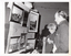 B&W photo of two people looking at several paintings hanging on a display board. In the background, there is another person standing in front of a table full of potted plants and other items.
