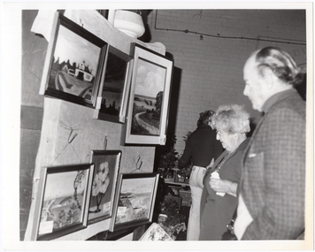 B&W photo of two people looking at several paintings hanging on a display board. In the background, there is another person standing in front of a table full of potted plants and other items.