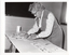 B&W photo of a woman leaning over a trestle table as she arranges some cards for display.