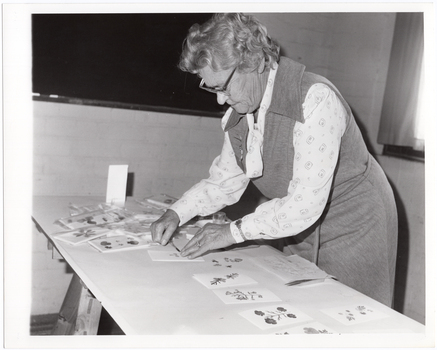 B&W photo of a woman leaning over a trestle table as she arranges some cards for display.
