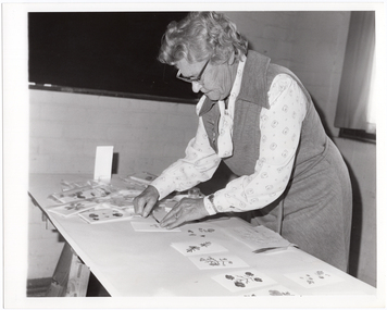 B&W photo of a woman leaning over a trestle table as she arranges some cards for display.