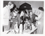 B&W photo of a several people, of all ages, browsing at a bric-a-brac stall. In front of them is a trestle table full of various items, from vases and small dishes to a vacuum drink flask and some knitted or crocheted items.