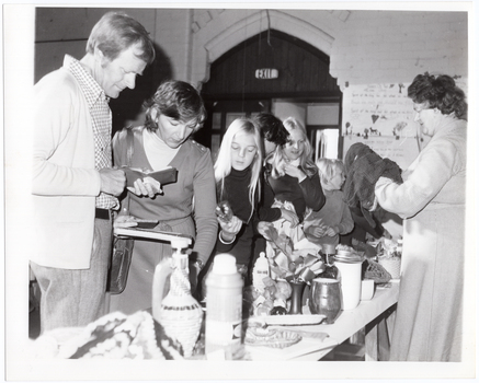 B&W photo of a several people, of all ages, browsing at a bric-a-brac stall. In front of them is a trestle table full of various items, from vases and small dishes to a vacuum drink flask and some knitted or crocheted items.