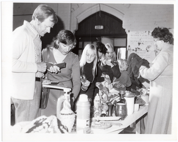B&W photo of a several people, of all ages, browsing at a bric-a-brac stall. In front of them is a trestle table full of various items, from vases and small dishes to a vacuum drink flask and some knitted or crocheted items.