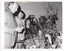 B&W photo of two people, standing. They are looking down at a table full of potted plants of different varieties and sizes.