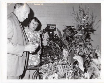 B&W photo of two people, standing. They are looking down at a table full of potted plants of different varieties and sizes.