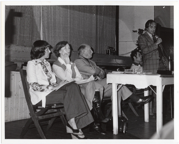 B&W photo of a man, standing, speaking into a microphone on a stand. He is wearing a checked suit, with a shirt and tie. Behind him, there are four people seated on folding wooden bench seats. There is a small painted wooden table in front of them, on which there is a video player and various pamphlets. Behind them is the edge of a stage, with a large stage curtain/backdrop in the background. There is a radio/cassette player and a large potted plant in the front corner of the stage.