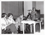 B&W photo of a man, standing, speaking into a microphone on a stand. He is wearing a checked suit, with a shirt and tie. Behind him, there are four people seated on folding wooden bench seats. There is a small painted wooden table in front of them, on which there is a video player and various pamphlets. Behind them is the edge of a stage, with a large stage curtain/backdrop in the background. There is a large potted plant in the front corner of the stage. There is another person (partially obscured) visible on the right edge of the frame.