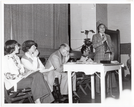 B&W photo of a man, standing, speaking into a microphone on a stand. He is wearing a checked suit, with a shirt and tie. Behind him, there are four people seated on folding wooden bench seats. There is a small painted wooden table in front of them, on which there is a video player and various pamphlets. Behind them is the edge of a stage, with a large stage curtain/backdrop in the background. There is a large potted plant in the front corner of the stage. There is another person (partially obscured) visible on the right edge of the frame.