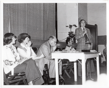 B&W photo of a man, standing, speaking into a microphone on a stand. He is wearing a checked suit, with a shirt and tie. Behind him, there are four people seated on folding wooden bench seats. There is a small painted wooden table in front of them, on which there is a video player and various pamphlets. Behind them is the edge of a stage, with a large stage curtain/backdrop in the background. There is a large potted plant in the front corner of the stage. There is another person (partially obscured) visible on the right edge of the frame.