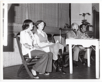 B&W photo of four people seated on some folding wooden bench seats. A man, sitting on one of the bench seats, is speaking into a microphone on a stand, which is situated in the gap between the two wooden bench seats. There is a small painted wooden table in front of them, on which there are various pamphlets. Behind them is the edge of a stage, with a large stage curtain/backdrop in the background. There is a radio/cassette player and a large potted plant in the front corner of the stage.