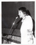 B&W photo of a woman speaking into a microphone on a stand. She is wearing a long-sleeved floral shirt under a plain short-sleeved jacket. The photo has been taken in three quarter profile. In the background, the front edge of a stage is visible with a stage curtain/backdrop behind it.