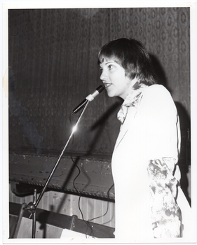 B&W photo of a woman speaking into a microphone on a stand. She is wearing a long-sleeved floral shirt under a plain short-sleeved jacket. The photo has been taken in three quarter profile. In the background, the front edge of a stage is visible with a stage curtain/backdrop behind it.