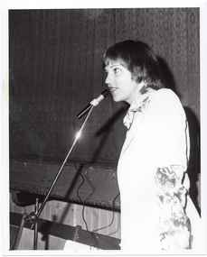 B&W photo of a woman speaking into a microphone on a stand. She is wearing a long-sleeved floral shirt under a plain short-sleeved jacket. The photo has been taken in three quarter profile. In the background, the front edge of a stage is visible with a stage curtain/backdrop behind it.