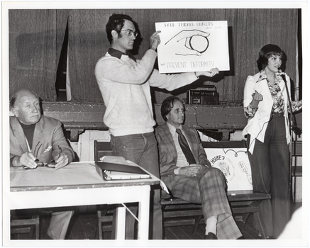 B&W photo of a woman, standing, speaking into a microphone on a stand. She is wearing a long-sleeved floral shirt under a pale short-sleeved jacket, and trousers. There is a wooden folding bench seat beside her. Two people are sitting on the bench seat while, between them, there is a man standing in front of it. He is holding up a sign which depicts a sketch drawing of a hand holding a spherical object and text that says, "Hold thicker objects where possible and prevent deformity". In front of them, there is a small table with a folder on top. There is another sign (partially obscured) on the bench seat. Behind them is the edge of a stage, with a large stage curtain/backdrop in the background. There is a radio/cassette player on the stage.