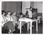 B&W photo of a man, standing, speaking into a microphone on a stand. He is wearing eye glasses, and a dark shirt over a light-coloured jumper. Beside him, there are four people seated on folding wooden bench seats. There is a small painted wooden table in front of them, on which there is a video player and various pamphlets. Behind them is the edge of a stage, with a large stage curtain/backdrop in the background. To the right of the frame, there is another woman sitting on another folding wooden bench seat.