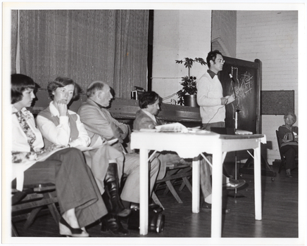 B&W photo of a man, standing, speaking into a microphone on a stand. He is wearing eye glasses, and a dark shirt over a light-coloured jumper. Beside him, there are four people seated on folding wooden bench seats. There is a small painted wooden table in front of them, on which there is a video player and various pamphlets. Behind them is the edge of a stage, with a large stage curtain/backdrop in the background. To the right of the frame, there is another woman sitting on another folding wooden bench seat.