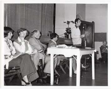 B&W photo of a man, standing, speaking into a microphone on a stand. He is wearing eye glasses, and a dark shirt over a light-coloured jumper. Beside him, there are four people seated on folding wooden bench seats. There is a small painted wooden table in front of them, on which there is a video player and various pamphlets. Behind them is the edge of a stage, with a large stage curtain/backdrop in the background. To the right of the frame, there is another woman sitting on another folding wooden bench seat.