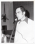 B&W close-up photo of a man, standing, speaking into a microphone on a stand. He is wearing eye glasses, and a dark shirt under a pale woollen jumper. In the background, there is a seated person (partially obscured). Behind them is the edge of a stage, with a large stage curtain/backdrop in the background. In the front corner of the stage, there is a large public address speaker on the floor and a potted plant inside a macrame holder hanging from the wall or ceiling.
