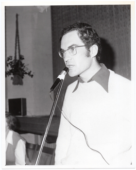 B&W close-up photo of a man, standing, speaking into a microphone on a stand. He is wearing eye glasses, and a dark shirt under a pale woollen jumper. In the background, there is a seated person (partially obscured). Behind them is the edge of a stage, with a large stage curtain/backdrop in the background. In the front corner of the stage, there is a large public address speaker on the floor and a potted plant inside a macrame holder hanging from the wall or ceiling.