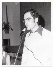 B&W close-up photo of a man, standing, speaking into a microphone on a stand. He is wearing eye glasses, and a dark shirt under a pale woollen jumper. In the background, there is a seated person (partially obscured). Behind them is the edge of a stage, with a large stage curtain/backdrop in the background. In the front corner of the stage, there is a large public address speaker on the floor and a potted plant inside a macrame holder hanging from the wall or ceiling.