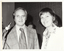B&W close up photo of two people posing for a Photograph. Both the man and the woman are smiling. The man is standing behind a microphone on a stand. He is wearing a checked jacket with a shirt and tie. The woman is wearing a long-sleeved floral shirt under a plain jacket. There is a third person (partially obscurred) in the background.