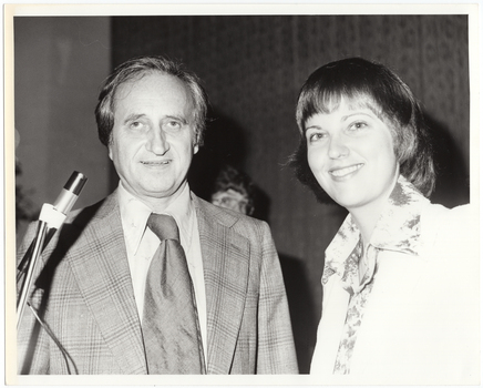 B&W close up photo of two people posing for a Photograph. Both the man and the woman are smiling. The man is standing behind a microphone on a stand. He is wearing a checked jacket with a shirt and tie. The woman is wearing a long-sleeved floral shirt under a plain jacket. There is a third person (partially obscurred) in the background.