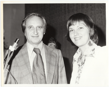 B&W close up photo of two people posing for a Photograph. Both the man and the woman are smiling. The man is standing behind a microphone on a stand. He is wearing a checked jacket with a shirt and tie. The woman is wearing a long-sleeved floral shirt under a plain jacket. There is a third person (partially obscurred) in the background.