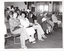 B&W photo of a large group of people seated on rows of chairs in a hall. Two women in the front row are looking directly at the camera, while many others are looking down at the program guide which they are holding in their hands. In the background, a few people can be seen standing at the back of the hall and near a set of glass double doors or windows. There is a lot of bright light coming in through the glass doors/windows.