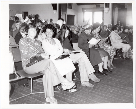 B&W photo of a large group of people seated on rows of chairs in a hall. Two women in the front row are looking directly at the camera, while many others are looking down at the program guide which they are holding in their hands. In the background, a few people can be seen standing at the back of the hall and near a set of glass double doors or windows. There is a lot of bright light coming in through the glass doors/windows.