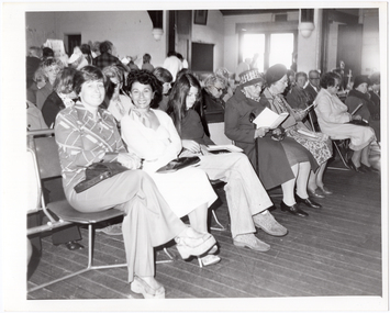 B&W photo of a large group of people seated on rows of chairs in a hall. Two women in the front row are looking directly at the camera, while many others are looking down at the program guide which they are holding in their hands. In the background, a few people can be seen standing at the back of the hall and near a set of glass double doors or windows. There is a lot of bright light coming in through the glass doors/windows.