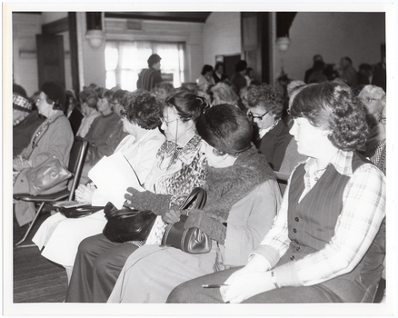 B&W photo of a large group of people seated on rows of chairs in a hall. Many people are looking across to one side of the hall. In the background, a few people can be seen standing at the back of the hall. There is a large set of windows, partially obscured by the figure of a person walking towards the back of the hall.